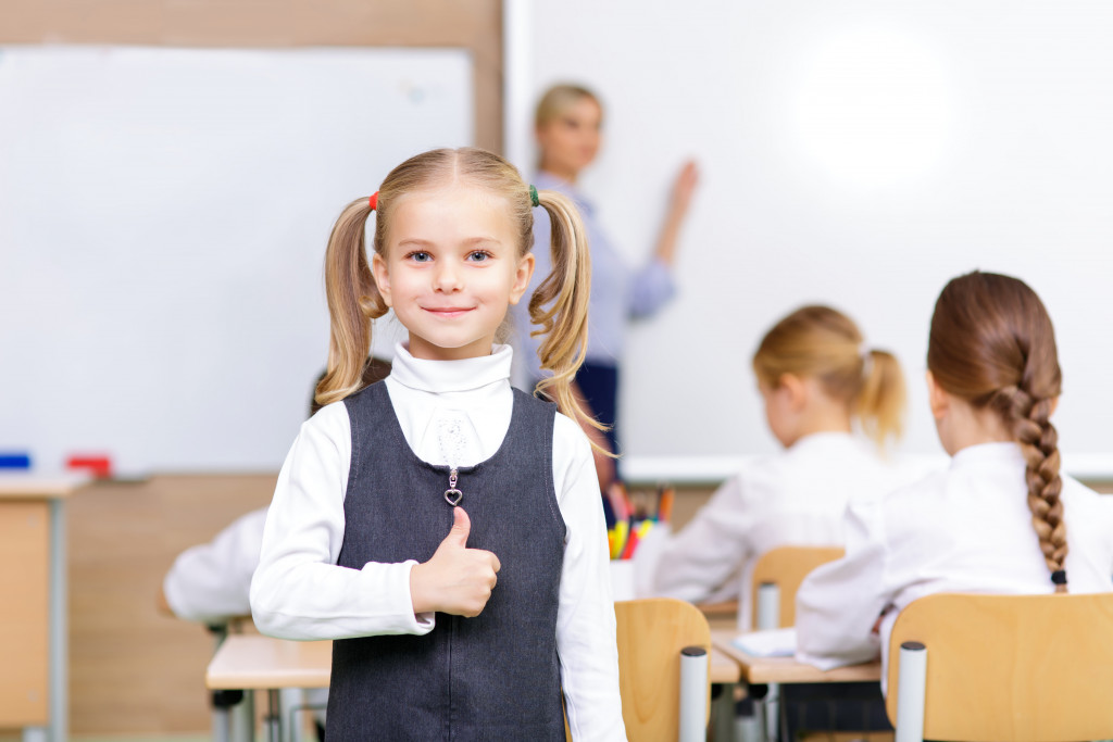 Young girl giving a thumbs up sign with the teacher and class in the background.